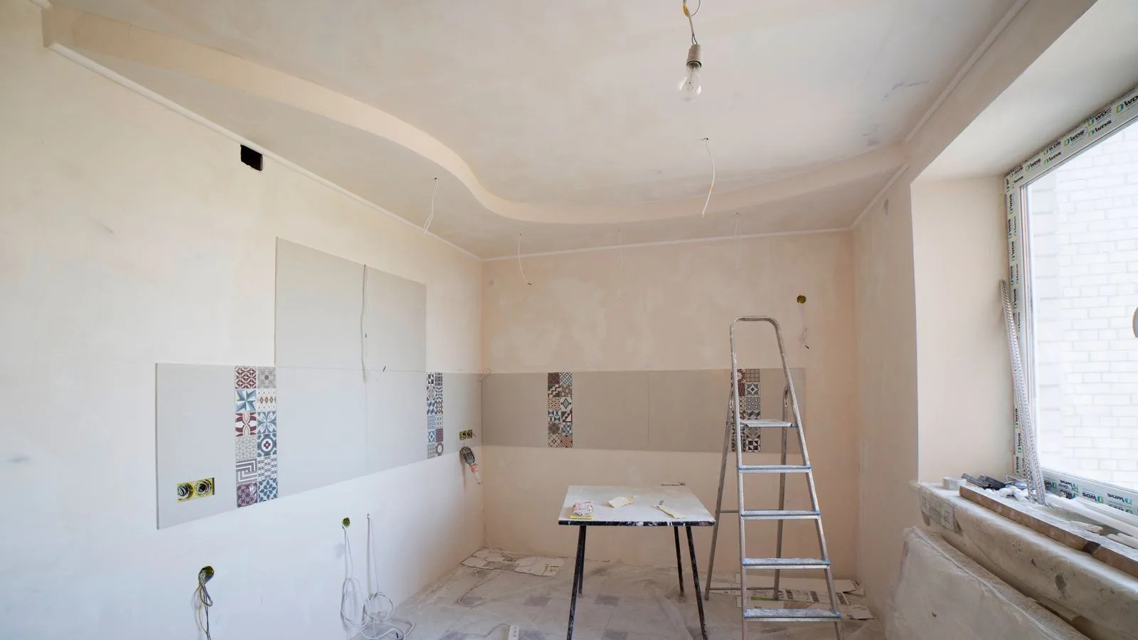 Kitchen mid-remodel with patterned tile going up, stepladder, drop sheets and exposed wiring